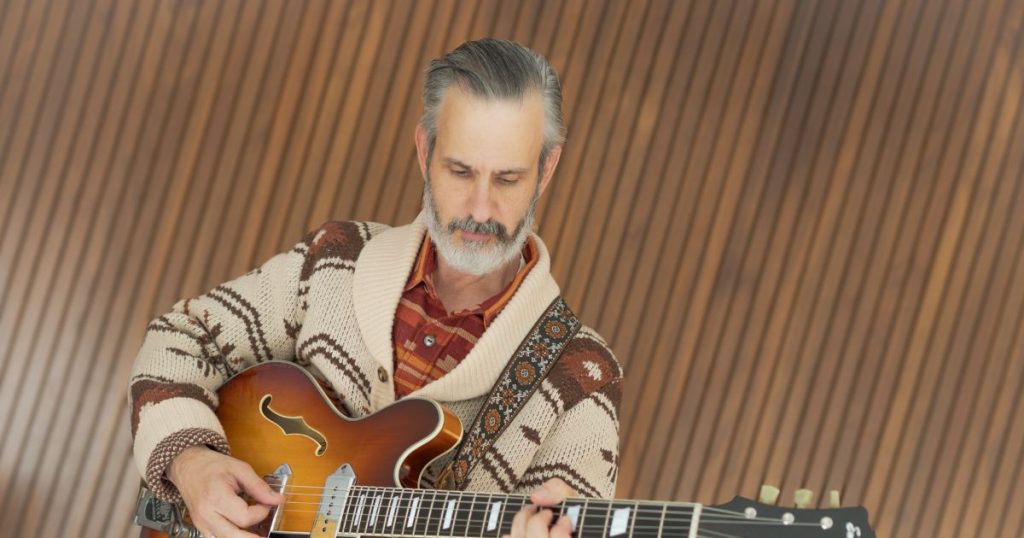 Wayne McRae playing a sunburst hollowbody guitar in front of a warm wood-paneled wall.